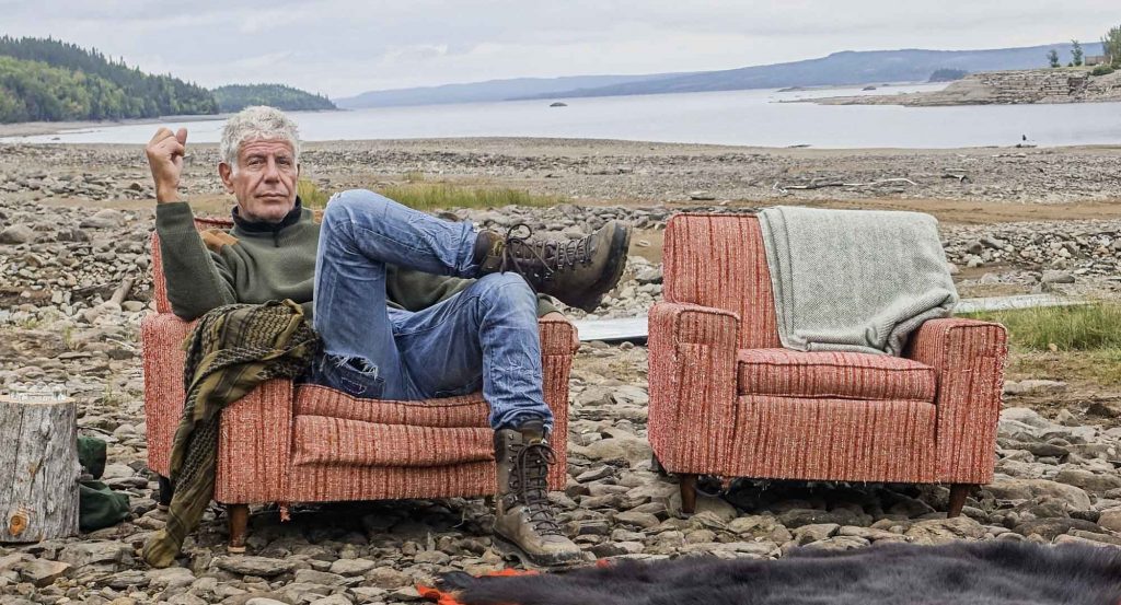 Anthony Bourdain in Newfoundland for his show Parts Unknown sitting on a rocky beach in an old chair with a bear rug in front of him and another identical empty chair beside him. 